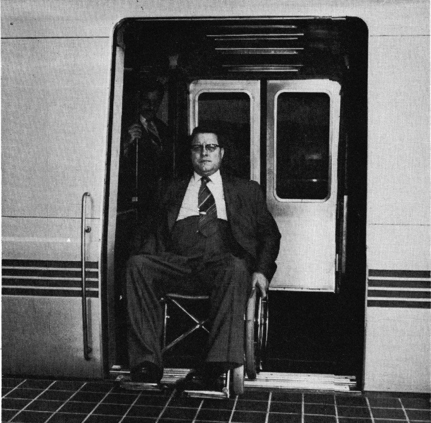 Photo of harold Wilson in his wheelchair, seated in the doorway of a train. He wears a suit and tie and glasses.