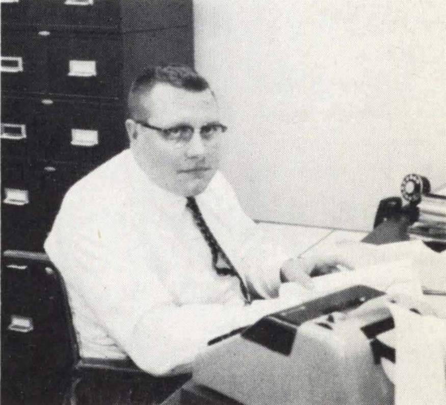 A black and white photo of Harold Willson at a desk with papers and a typewriter. He wears a tie and glasses and white buttondown.