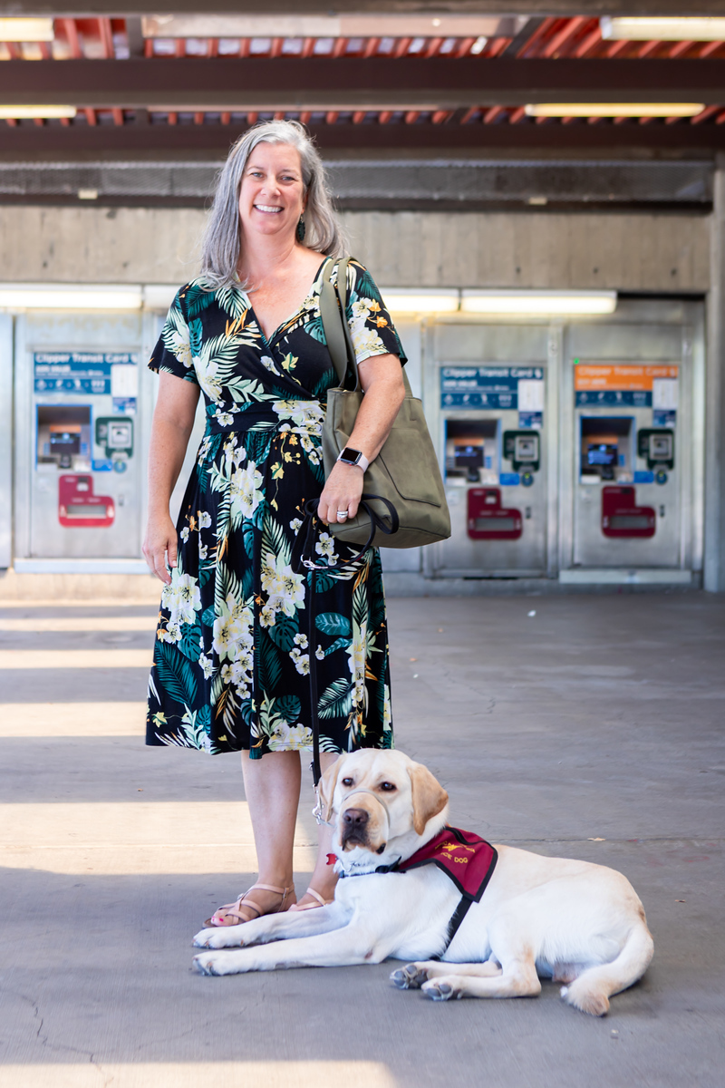 Puppy trainer Suzie Scher with trainee service dog Sheldon standing in the unpaid area of Concord Station. Sheldon is laying out her feet.