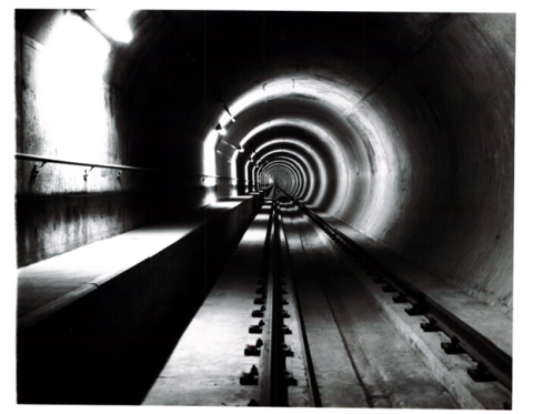 Black and white photograph of an empty, curved Transbay Tube with tracks extending into the distance, illuminated by overhead lights.