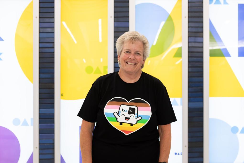 Terri Hodges smiling in a new BART Pride shirt in front of bold yellow and blue shapes.