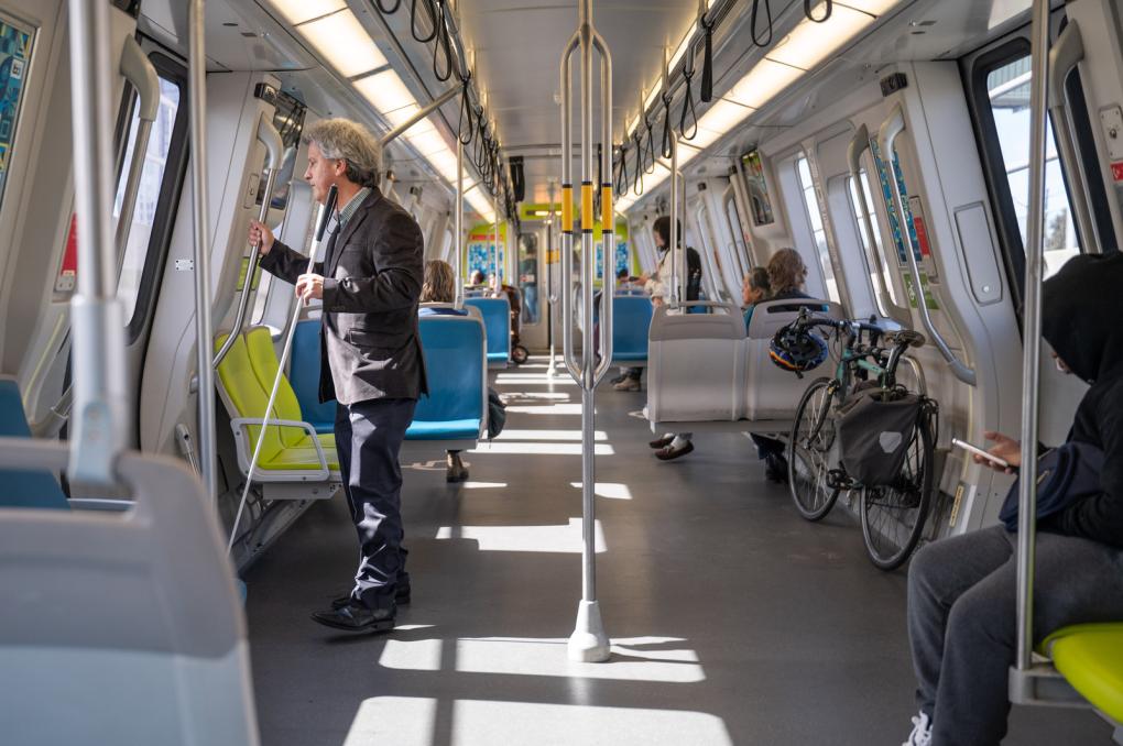 Interior of a BART car with passengers in various seats and one standing holding onto a pole. The carriage features blue and green seats, metal poles for stability, and clear windows showing daylight outside. A bicycle is parked in a designated area. Text or symbols on the train are not clearly visible.