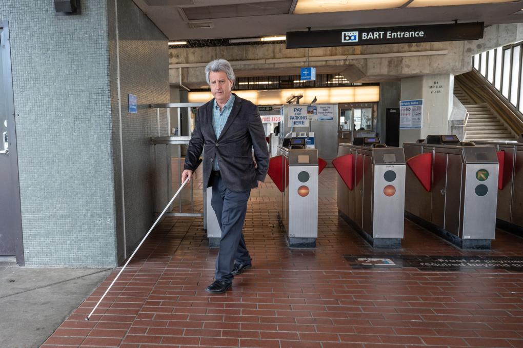 A person using a white cane exits a BART (Bay Area Rapid Transit) station entrance, navigating through the ticket barrier in an urban setting.