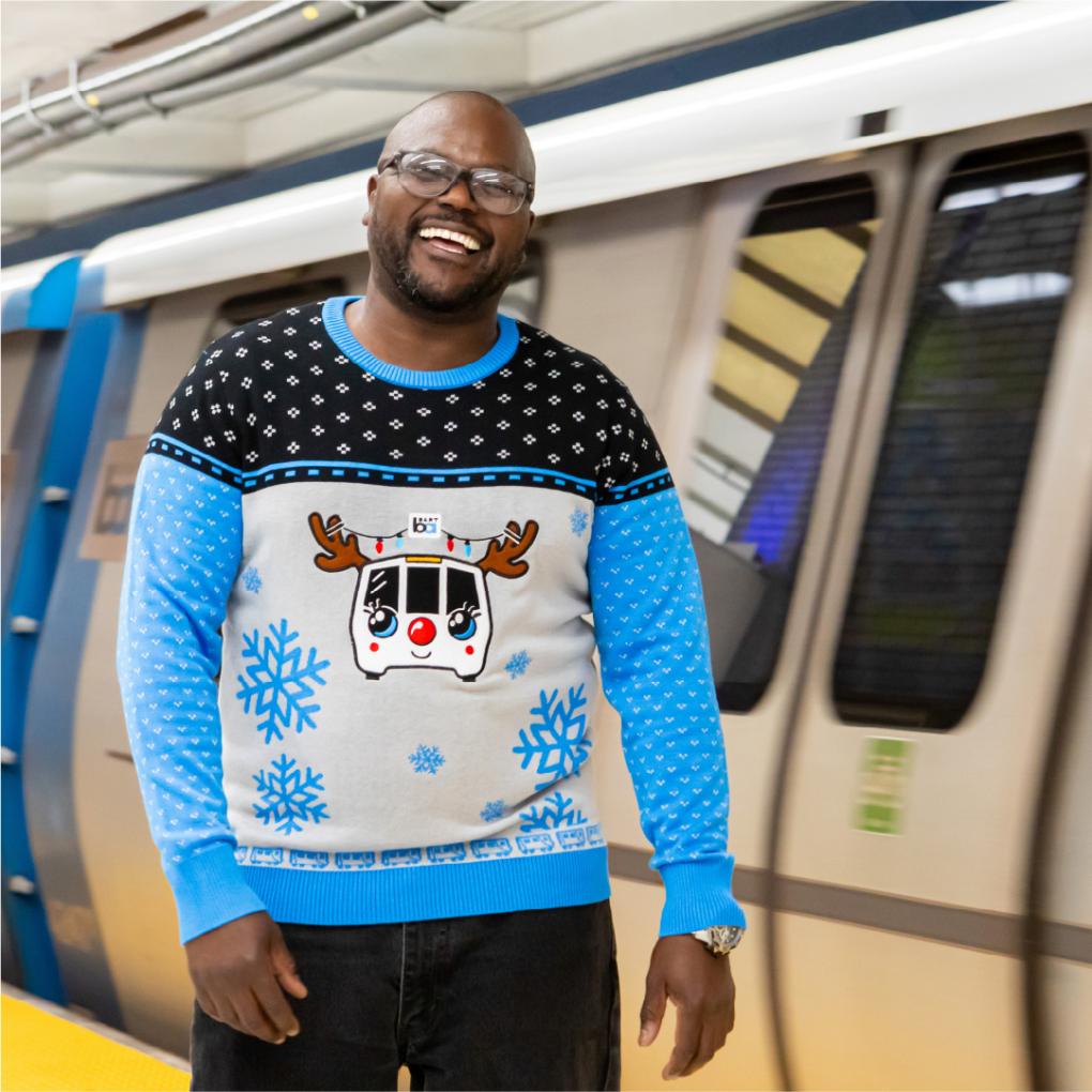 Person smiling in a BART holiday sweater in front of a BART train 