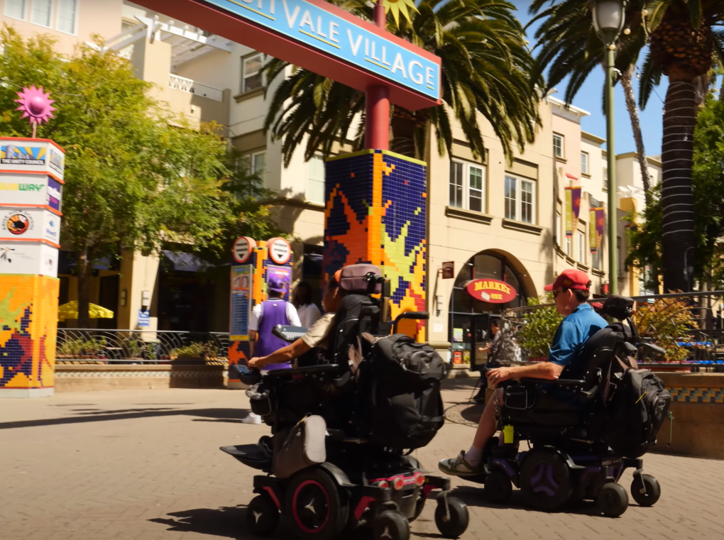 Two individuals in motorized wheelchairs move through a vibrant outdoor shopping area beside Fruitvale Station with palm trees, under a sign that reads "Fruitvale Village."