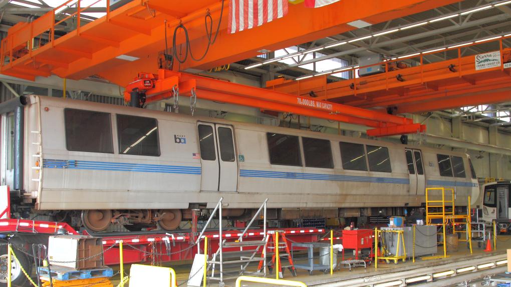 A BART train in a maintenance facility with overhead cranes and various equipment around.