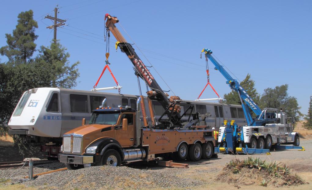 BART train car being lifted by two cranes onto a transport trailer in a dry, grassy field with power lines and clear skies in the background.