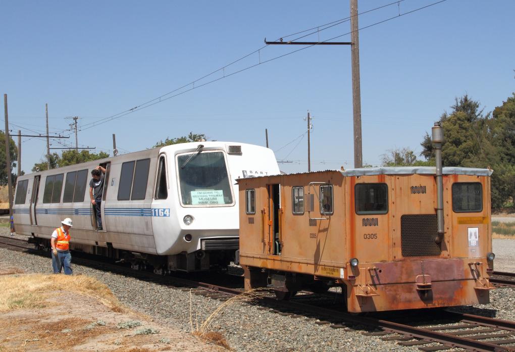 A BART train on the left with a worker standing by, and an maintenance vehicle on the right, both on tracks under a clear sky.