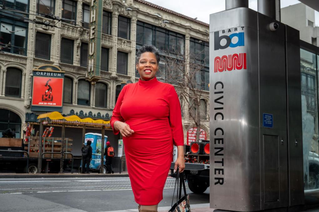 Sherri Young pictured next to a Civic Center BART and Muni sign in front of the Orpheum Theater. She is wearing a red dress and carries a purse