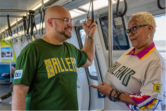 Two adults are conversing and smiling inside a train. One person is wearing a green shirt with "Ballers" printed on it, and the other is in a colorful top with a sports jersey design that says "Oakland". They are standing and holding onto handrails.