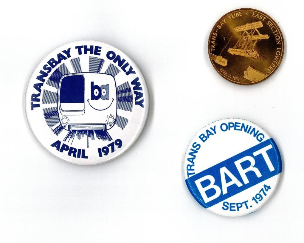 Three commemorative BART pins: one with a cartoon train and the text "Transbay the Only Way April 1979," another with a spacecraft design and the text "Man's Last Hope - Eastbound," and a third with the text "Trans Bay Opening BART Sept 1974"