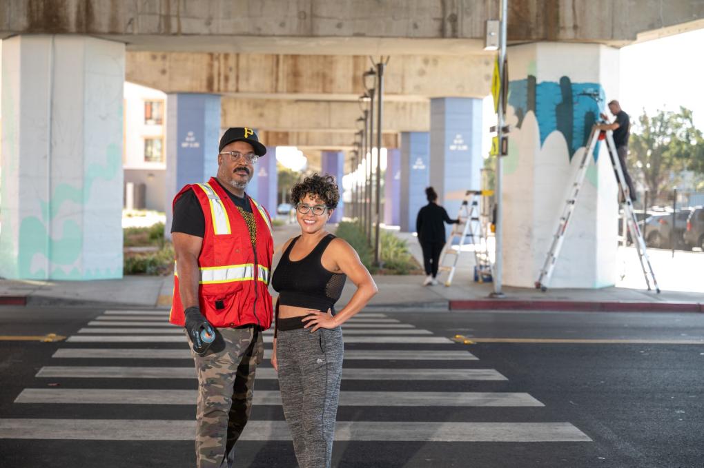 Artists Refa1 and Favianna Rodriguez smiling at the camera, standing at a zebra crossing under an overpass with mural painting activity visible in the background. 