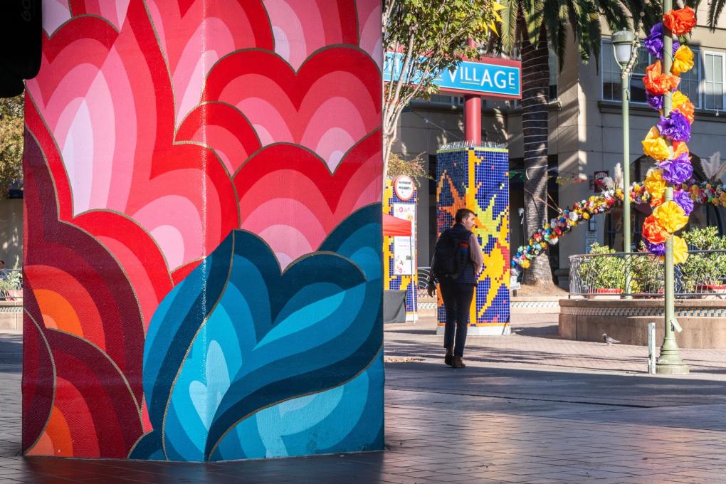 Colorful mural featuring large flower designs on the left, with a person walking by. On the right, a decorated area labeled "Fruitvale Village" with festive orange and purple flower arrangements and a brightly colored star-shaped figure.